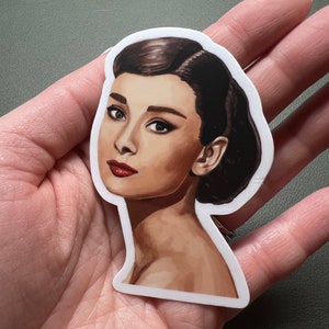 以下が含まれることがあります： 白黒のイラストスタイルで描かれたオードリー・ヘップバーンの肖像画が描かれたステッカー。ステッカーは白い背景です。