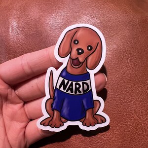May include: A sticker featuring a cartoon dog with brown fur and floppy ears. The dog is wearing a blue sweater with the word "NARD" in white letters. The dog is smiling with its tongue out.
