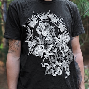 Op de afbeelding: Zwart T-shirt met een witte grafische print van een wolf met een slang eromheen gewikkeld. De wolf huilt en de slang bijt de wolf in de nek. De afbeelding is omgeven door een ster.