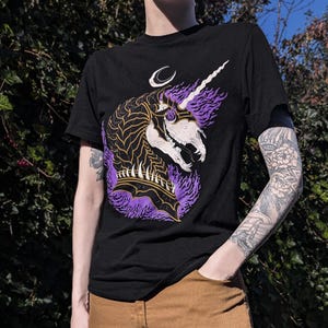 Peut inclure: T-shirt noir avec un imprimé graphique d'une tête de licorne violette et dorée avec une lune croissante au-dessus.