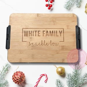 May include: A rectangular bamboo snack box with black handles, engraved with "WHITE FAMILY snackle box." The box is surrounded by Christmas decorations, including ornaments, pine branches, and berries.