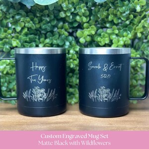 May include: Two matte black mugs with silver-toned rims and handles. The left mug is engraved with "Happy Ten Years" and a wildflower design. The right mug is engraved with "Sarah & Emmett" and "5-12-19" above a matching wildflower design.