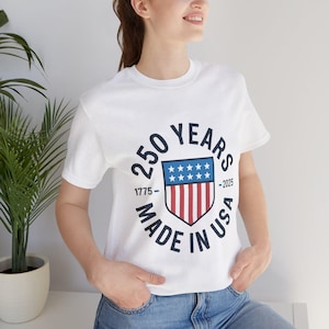 May include: White t-shirt featuring a graphic commemorating 250 years of the USA. The design includes a shield with stars and stripes, the years 1775-2025, and the text "Made in USA."