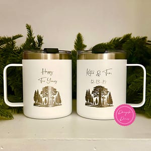 May include: Two white stainless steel mugs with black lids and handles. The left mug features a forest graphic and the text "Happy Ten Years". The right mug features a forest graphic and the text "Katie & Tim 12-13-14". A pink circular logo is present.