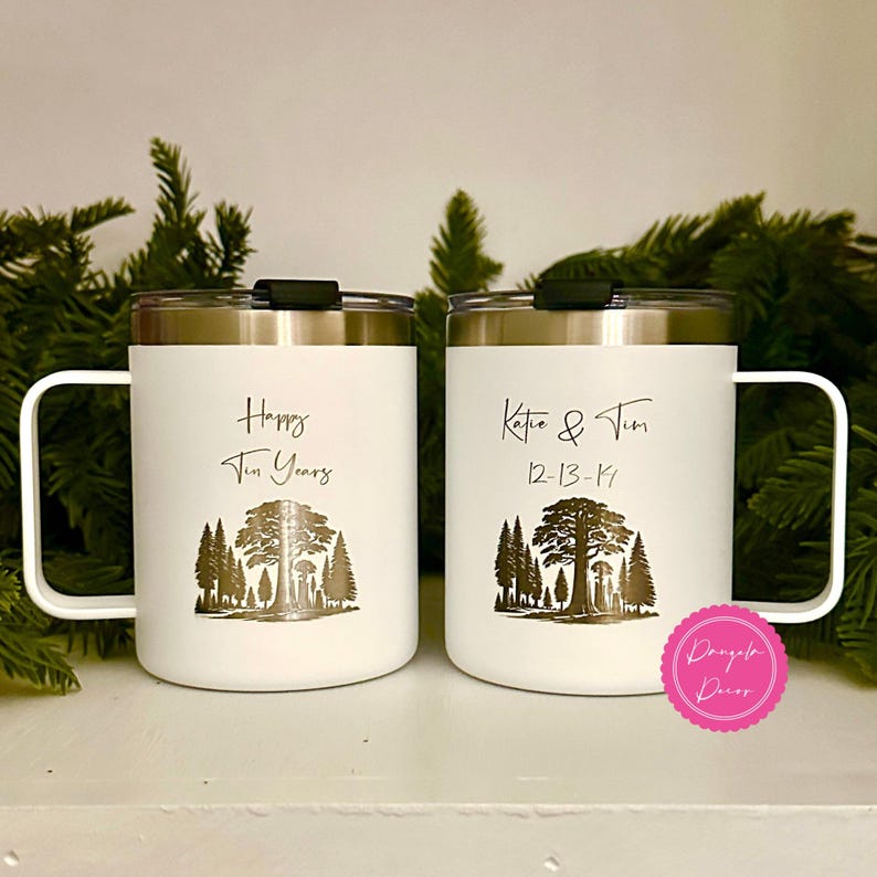 May include: Two white stainless steel mugs with black lids and handles. The left mug features a forest graphic and the text "Happy Ten Years". The right mug features a forest graphic and the text "Katie & Tim 12-13-14". A pink circular logo is present.