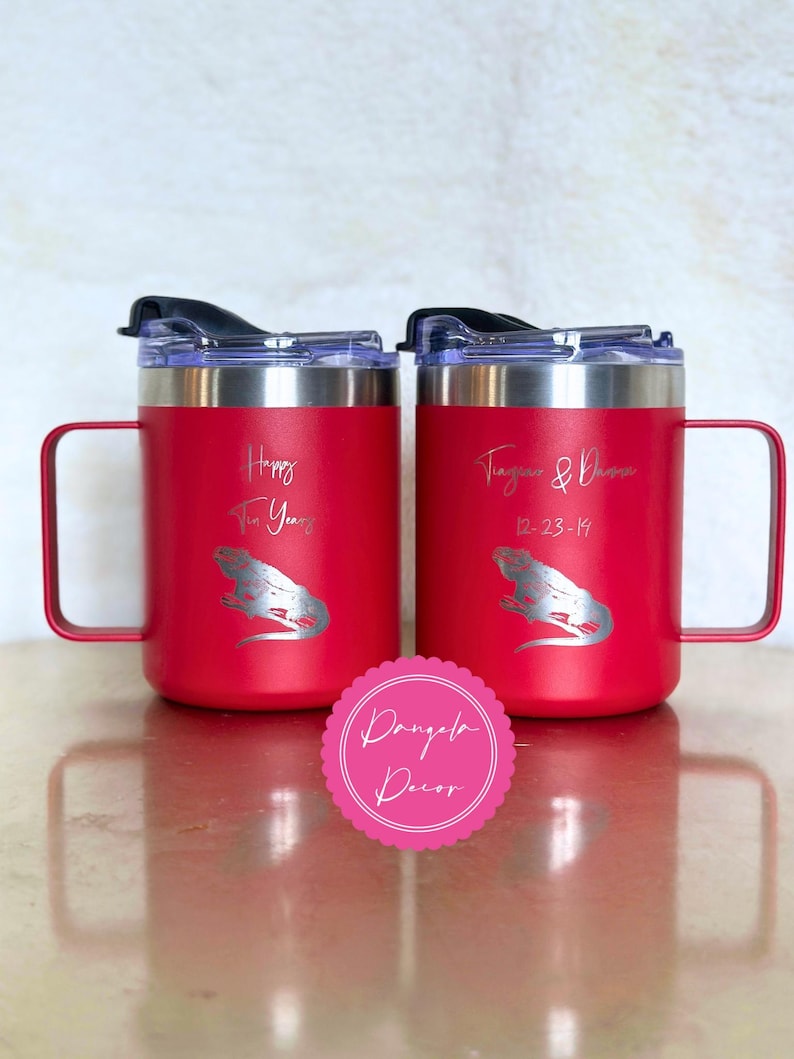 May include: Two red stainless steel mugs with handles and clear lids. The left mug features the text Happy Ten Years and an iguana graphic. The right mug features the text Tiara & Damon 12-23-14 and an iguana graphic. A pink circular logo reads Dangela Decor.