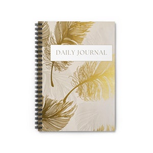 May include: A spiral-bound daily journal with a cream-colored cover featuring gold feather illustrations. The words "DAILY JOURNAL" are printed in a white rectangular box across the top of the journal.