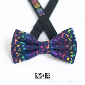 May include: A navy blue bow tie with a rainbow musical note pattern. The bow tie is tied with black adjustable straps that say "Guys + Ties".