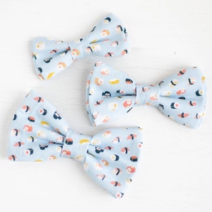 May include: Three blue bow ties with a sushi print pattern. The bow ties are arranged in a triangle shape on a white surface.