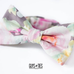 May include: A light pink bow tie with a floral pattern in shades of pink, green, and yellow. The bow tie is made of a soft, lightweight fabric and has a subtle sheen.  "GUYS + TIES" is printed in black text on the white background.