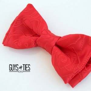 May include: A red bow tie with a patterned design. The bow tie is made of a soft, textured fabric. The bow tie is tied in a classic bow shape. The bow tie is a great accessory for any formal occasion.
