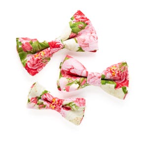 May include: Three floral bow ties in different sizes. The bow ties are made of a white fabric with a pink and green floral pattern.