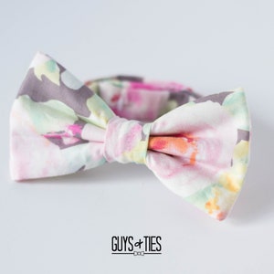 May include: A bow tie with a floral pattern in shades of pink, green, and gray. The bow tie is made of fabric and has a white background.