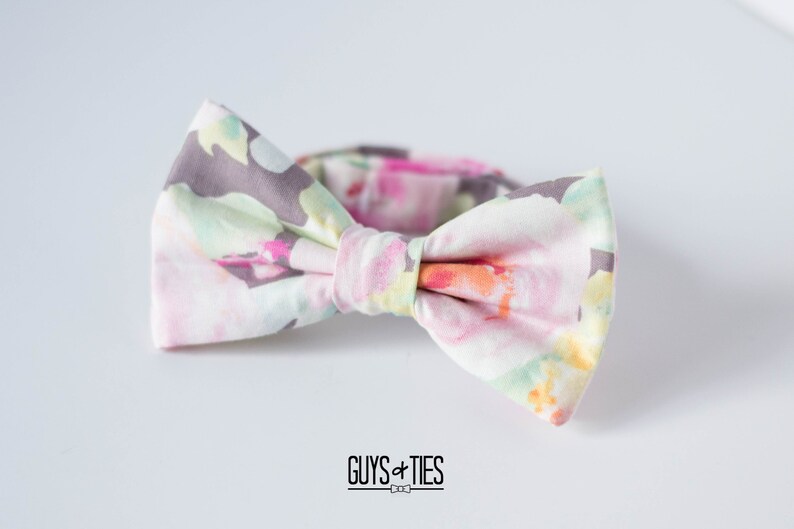 May include: A bow tie with a floral pattern in shades of pink, green, and gray. The bow tie is made of fabric and has a white background.