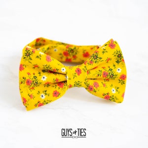 May include: A yellow bow tie with a floral pattern featuring red and white flowers. The bow tie is made of fabric and has a classic bow shape.