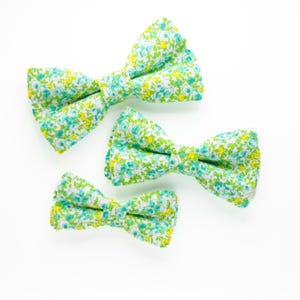 May include: Three floral bow ties in different sizes. The bow ties are made of a white fabric with a green and yellow floral pattern.