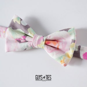 May include: A floral bow tie with a light pink, yellow, and green pattern on a white background. The bow tie has a gray adjustable strap.