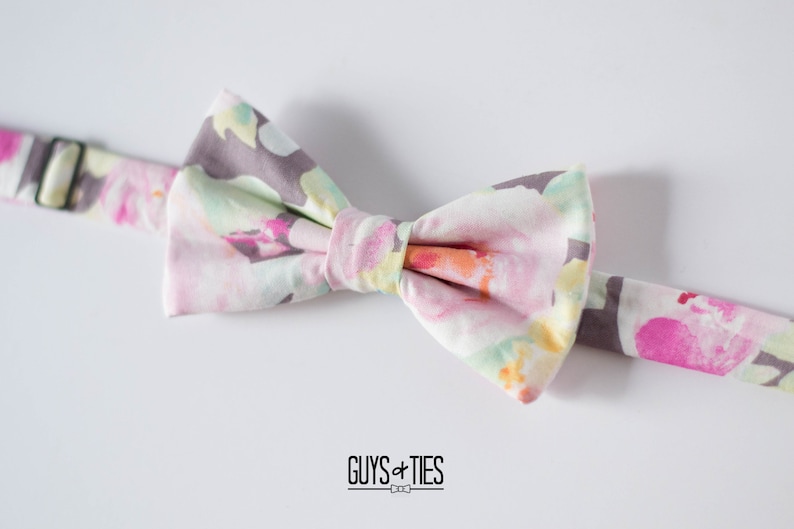 May include: A floral bow tie with a light pink, yellow, and green pattern on a white background. The bow tie has a gray adjustable strap.
