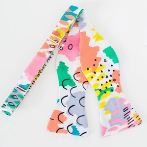 May include: A white bow tie with a colorful abstract pattern featuring pink, green, yellow, and blue. The pattern includes circles, squiggles, and wavy lines.