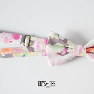 May include: A pink and white bow tie with a floral pattern. The bow tie is attached to a matching strap with the text "GUYS + TIES" printed on it.