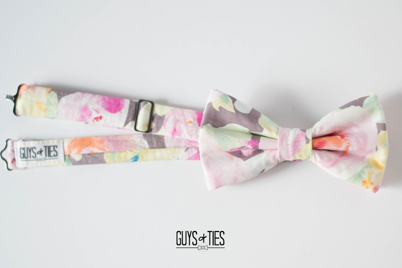 May include: A pink and white bow tie with a floral pattern. The bow tie is attached to a matching strap with the text "GUYS + TIES" printed on it.