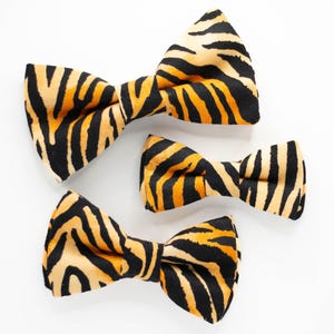 May include: Three bow ties in a tiger print pattern. The largest bow tie is orange and black, the medium bow tie is orange and black, and the smallest bow tie is orange and black.