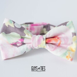 May include: A bow tie with a floral pattern in shades of pink, green, and gray. The bow tie is made of fabric and has a white background.