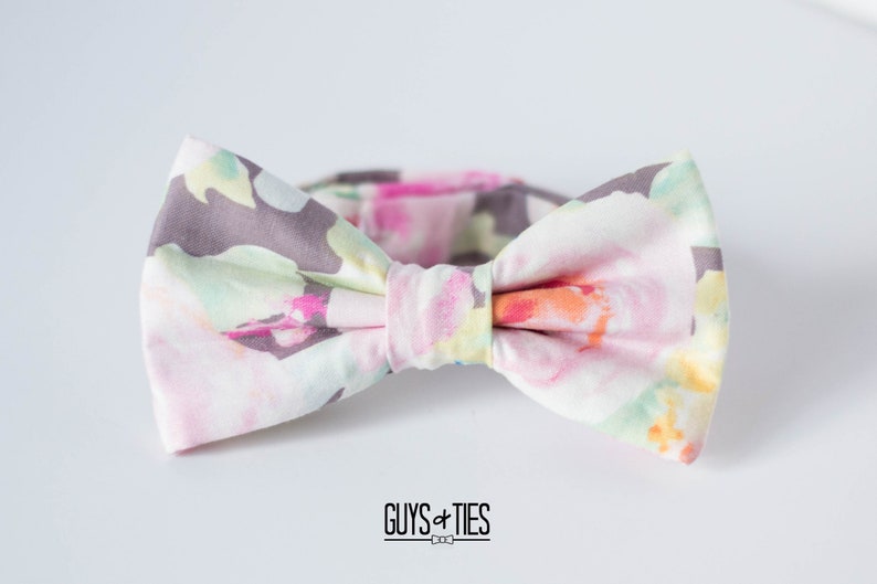 May include: A bow tie with a floral pattern in shades of pink, green, and gray. The bow tie is made of fabric and has a white background.