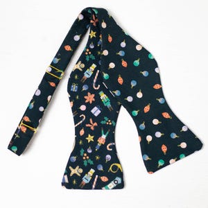 May include: A dark green bow tie featuring a Christmas-themed pattern with baubles, candy canes, stars, and nutcrackers. The adjustable strap has gold-coloured metal hardware. The bow tie is a self-tie style.