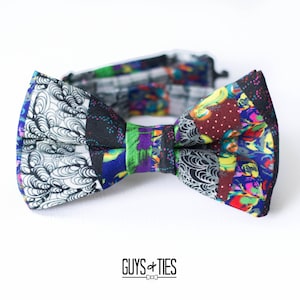 May include: A colourful bow tie with a patchwork design featuring black, white, blue, red, green and yellow patterns. The bow tie is made of fabric and has a textured surface.
