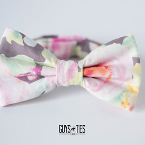 May include: A bow tie with a floral pattern in shades of pink, green, and yellow on a white background. The bow tie has a gray center.