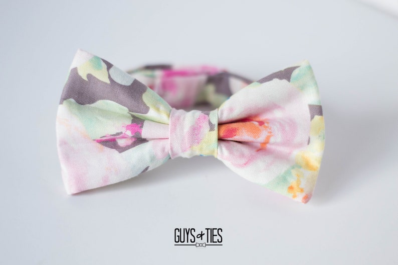 May include: A bow tie with a floral pattern in shades of pink, green, and yellow on a white background. The bow tie has a gray center.