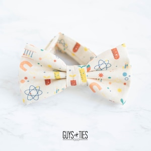 Könnte beinhalten: Ein beiger Fliege mit einem wissenschaftlichen Muster mit bunten Illustrationen von Atomen, Bechergläsern und anderen wissenschaftlichen Symbolen. Die Fliege ist aus einem weichen, bequemen Stoff gefertigt und perfekt, um jedem Outfit einen Hauch von Verspieltheit zu verleihen.