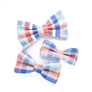 May include: Three plaid bow ties in different sizes. The largest bow tie is blue, pink, and white. The medium bow tie is blue, pink, and white. The smallest bow tie is blue, pink, and white.