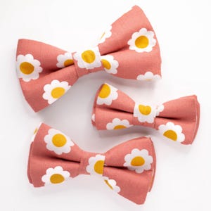 May include: Three pink bow ties with white daisy patterns. The bow ties are different sizes.