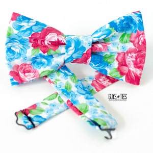 May include: A floral bow tie with a white background and blue and pink flowers. The bow tie is tied and ready to wear. The bow tie is made by Guys + Ties.