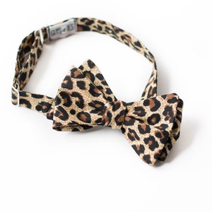 May include: A brown and black leopard print bow tie with an adjustable strap. The strap has the text "BUTS-TIES" printed on it.