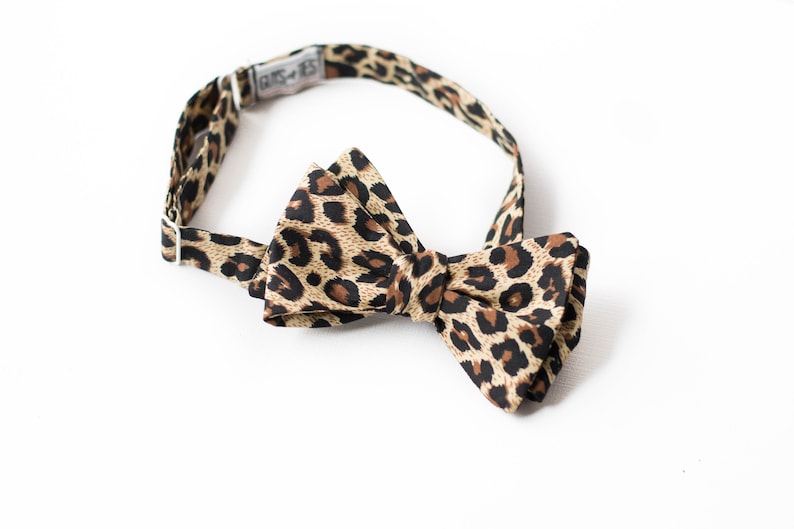 May include: A brown and black leopard print bow tie with an adjustable strap. The strap has the text "BUTS-TIES" printed on it.