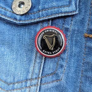 Puede incluir: Un pin redondo con el logotipo de Guinness, fondo negro y borde rojo, sujeto a una chaqueta vaquera azul. El pin dice "THE ORIGINAL EXTRA STOUT" y presenta un símbolo de arpa.
