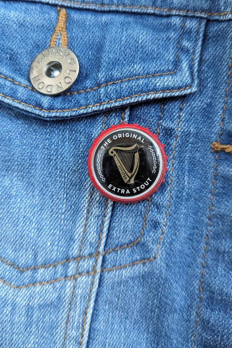 Pode incluir: Um alfinete redondo com o logotipo da Guinness, fundo preto e borda vermelha, preso a uma jaqueta jeans azul. O alfinete diz "THE ORIGINAL EXTRA STOUT" e apresenta um s&iacute;mbolo de harpa.