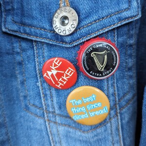 Pode incluir: Close-up de uma jaqueta jeans azul com um bot&atilde;o prateado e tr&ecirc;s pinos redondos. Os pinos apresentam o texto "TAKE HIKE!", "The best thing since sliced bread!" e um logotipo Guinness Extra Stout.