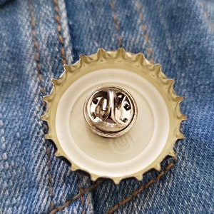 Pode incluir: Um alfinete de tampa de garrafa de metal com um pino prateado. O alfinete &eacute; preso ao centro da tampa da garrafa, que tem um interior branco e uma borda prateada e frisada. O fundo &eacute; um tecido de ganga azul.