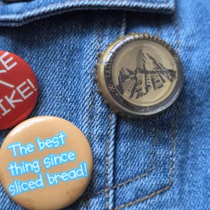 May include: Three circular pins attached to a blue denim fabric. One pin is red with white text, one is tan with blue text reading The best thing since sliced bread!, and one is a gold-colored metal bottle cap with a mountain graphic and text.
