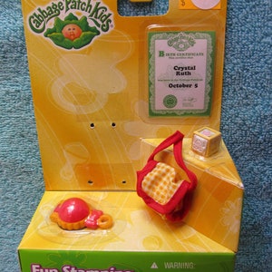 May include: A Cabbage Patch Kids Fun Stamping Babies playset with a yellow, green, and red color scheme. The set includes a red and yellow baby doll, a yellow and red diaper bag, and a yellow and red birth certificate. The birth certificate reads "This certifies that Crystal Ruth was born in the Cabbage Patch on October 5."