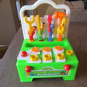 May include: A vintage Sesame Street workbench toy in green, white, and orange. The toy features Sesame Street characters holding tools, with shapes and pegs for hammering. The workbench has the text "Sesame Street Workbench".
