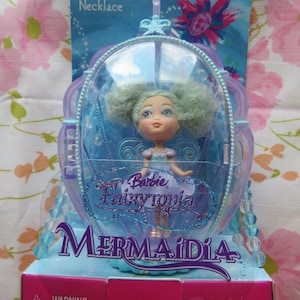 May include: A Barbie Fairytopia Mermaidia doll in a clear, blue, oval-shaped display case. The packaging reads "Sea Pixie Necklace" and "Barbie Fairytopia Mermaidia". The doll has green hair and a blue dress. The box is pink and blue.
