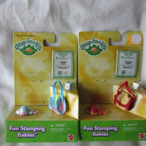 May include: Two Cabbage Patch Kids Fun Stamping Babies toy sets in yellow and green packaging. Each set includes a small doll accessory, a miniature handbag, and a stamp. The left set features Adelle Gail, and the right set features Crystal Rain.