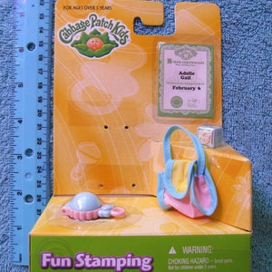 May include: A yellow, green, and blue Cabbage Patch Kids Fun Stamping Babies playset with a birth certificate, a small purse, and a rattle. The box has a warning label for choking hazards. The text on the box reads "Fun Stamping Babies" and "For Ages Over 3 Years".