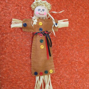 May include: A handmade scarecrow ornament with a straw hat and arms. The scarecrow has a painted face, a brown felt body, and is adorned with colorful buttons and raffia. A festive decoration for fall.