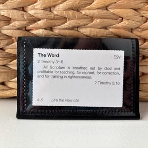 May include: Black leather card holder with a clear plastic window displaying a white card with the text "The Word" and a Bible verse from 2 Timothy 3:16 in the ESV translation. The card also includes the text "A-5 Live the New Life".